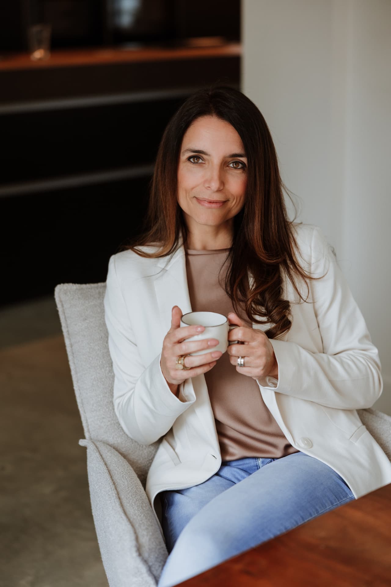 Joyce Schiffers zit lachend in een witte blazer met een kop koffie in haar handen