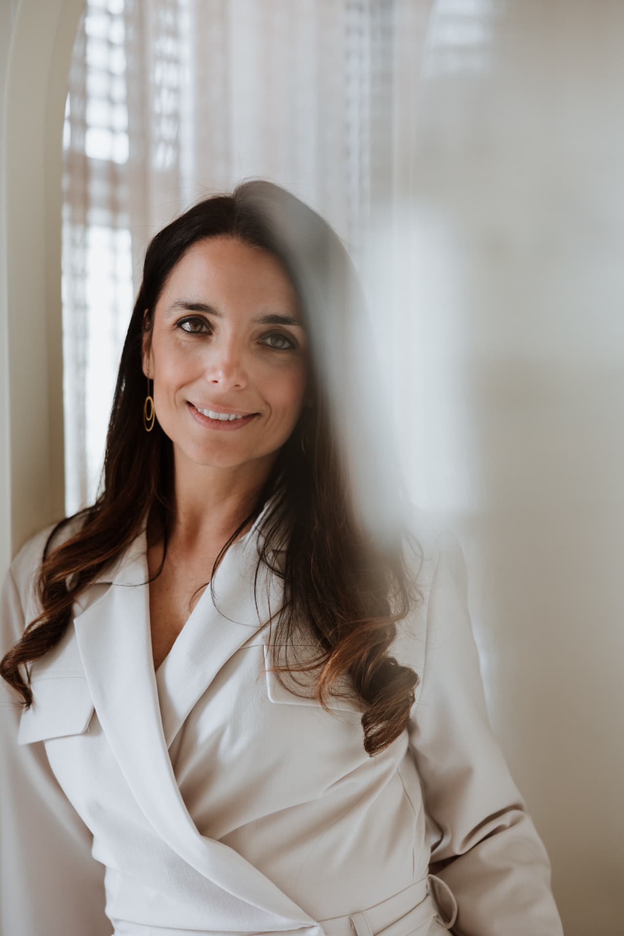 Joyce Schiffers in een crèmekleurige blazer, staand in een licht interieur met gordijnen